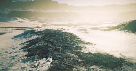 A vast landscape features snow covered terrain and rugged rocky formations illuminated by gentle sunlight. The scenery conveys tranquility and the beauty of nature at twilight.