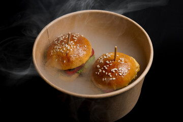 Two mini cheeseburgers with sesame buns are served in a cardboard bowl surrounded by smoky effects