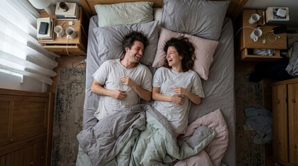 Fototapeta premium Top view of happy couple lying in bed laughing together. Cozy morning scene showing love and relationship intimacy. Ideal for lifestyle content and home comfort concepts.