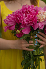 Close-up of a woman in a bright yellow dress holding a lush bouquet of vibrant pink and cream peonies, showcasing her light blue manicured nails and a fresh summer aesthetic.