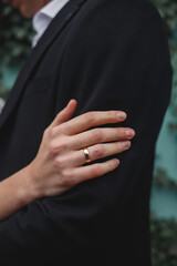 Elegant close-up of a hand wearing a gold wedding ring resting on a man's black suit sleeve, representing love, commitment, and marriage during a formal wedding ceremony.