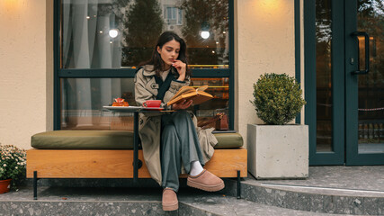 Young woman reading a book while enjoying coffee and pastry at an outdoor cafe