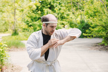 A focused man in a martial arts gi and headband practices outdoors amidst lush green foliage, showcasing dedication and a unique style.