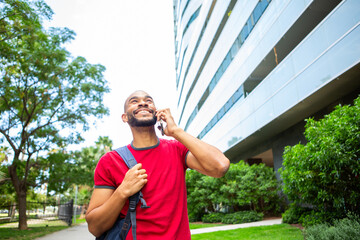 Smiling latin man in red shirt looking up while talking on phone outdoors near modern building
