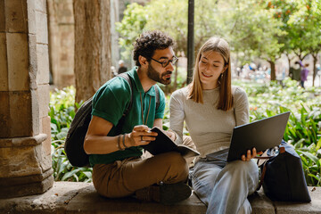 A man looks at a laptop that a woman is showing him, who is smiling and looking at a notebook he is holding while they sit on a wall