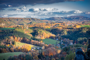 Saxon Switzerland, Bastei nature reserve. Panoramic view of a beautiful German village, surrounded by a vibrant autumn forest in the sunlight, mountains on the horizon, and a cloudy sky