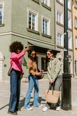 A woman is taking a picture on her phone while two female friends are standing next to her, looking at the phone that one of them is showing while one of them is leaning on a street lamp
