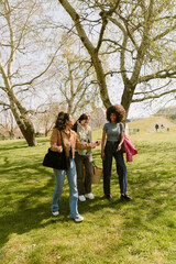 Fototapeta premium A group of three female friends walking on the grass carrying bags while laughing and one of them talking and holding a phone