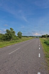 Two-lane asphalt rural road surrounded by green, bushes and trees, is sunny summer weather with a bright blue sky and some white clouds, Karuste, Saaremaa, Estonia.