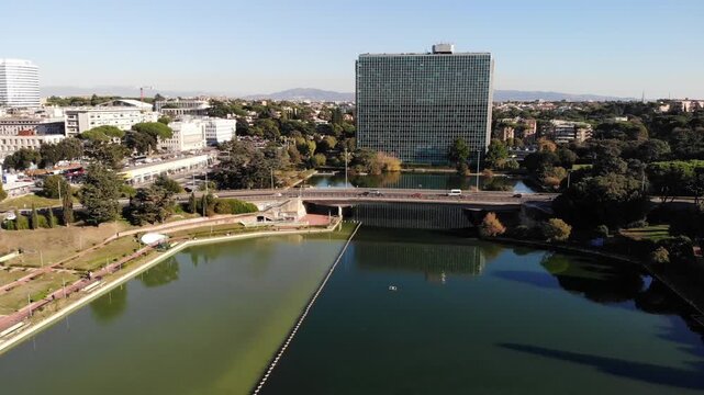 Forward aerial flight over EUR Park in Rome, showing water discharge in the lake, traffic crossing a three-lane bridge, and a modern building in the background.