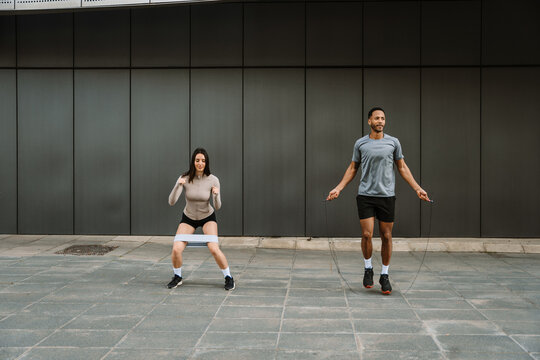 A male athlete jumps rope while a female athlete squats next to him with a rubber band on her legs
