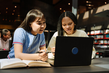 Two female students smiling and talking while looking at a laptop and sitting at a table while one of them holds a pen