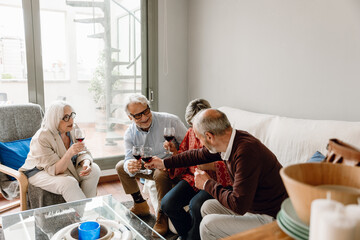 A group of four friends are talking and holding glasses while one of them is sitting on a chair and...
