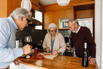 Two male friends holding glasses and watching their female friend cutting tomatoes while they stand at the table