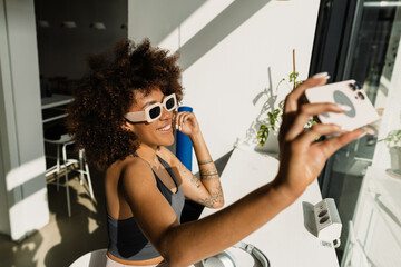 A woman sits at a table on a chair and smiles while holding a phone and posing in front of it