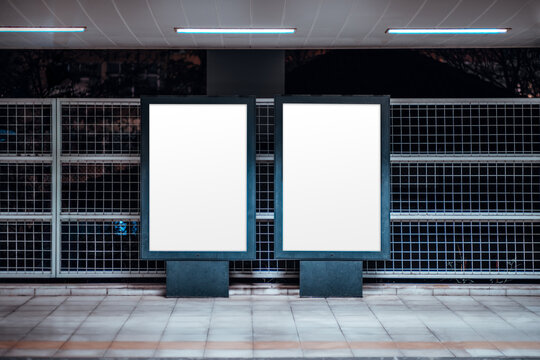 Urban mock-up scene with two blank vertical billboard posters templates at an outdoor subway platform, illuminated frames against metal grid wall, tiled floor and fluorescent lights