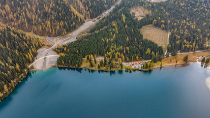 aerial landscape view of area along east coast of turquoise Antholzer See, a lake in the Antholzertal in South Tyrol, Italy, with "Antholzer Wildsee Haus" hotel, surrounded by autumn forest