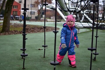 Obraz premium Toddler girl walking in rope climbing playground in autumn park, wearing warm colorful winter clothes. Outdoor activity for small child, balance and coordination development on modern playground.