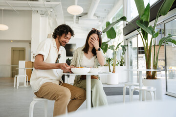 Obraz premium A man and a woman are laughing and sitting at a table while he holds cutlery in a plate and she covers her face with her hand