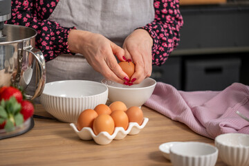 Female chef with manicured nails is cracking eggs into a white bowl while preparing ingredients on a wooden countertop, showcasing culinary skills and vibrant kitchen atmosphere