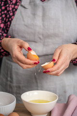 Female chef with pink nails is cracking an egg into a white bowl, wearing a gray apron, surrounded by fresh ingredients, showcasing culinary skills and kitchen creativity