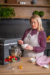 Woman in floral blouse and apron prepares dessert in modern kitchen, mixing ingredients in a bowl with fresh fruits on the counter, showcasing culinary creativity and home cooking