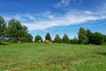 A picturesque summer landscape featuring hay bales, trees, and a bright blue sky.