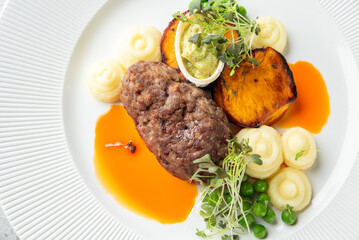 A gourmet meat patty served with white mashed potatoes, orange sweet potato slices, and green peas on a white plate with sauce