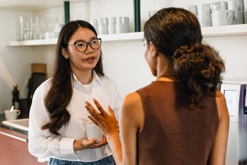 A female worker is holding a cup and talking to a female worker standing across from her and gesturing