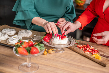 Two women, one in green and one in red, are decorating a dessert with fresh strawberries and flowers, showcasing a vibrant culinary experience in a cozy kitchen setting