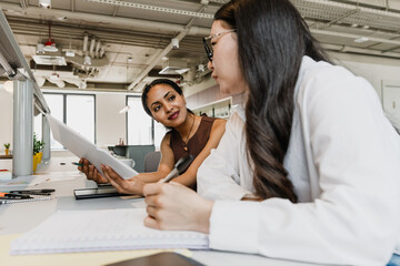 A female worker holds a folder and a pencil and smiles while listening to a female worker sitting next to her at a table and holding a pen