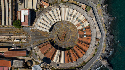 Perpendicular aerial view of an abandoned locomotive depot near the sea. An old industrial structure with a circular shape seen from above.