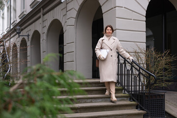 Woman in a stylish beige coat walks down stone steps, holding a white handbag, surrounded by greenery and elegant architecture, showcasing urban fashion and lifestyle