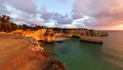 A dramatic tropical gorge at sunset, with limestone cliffs glowing warm orange over turquoise ocean waters and a secluded sandy beach under a colorful sky.