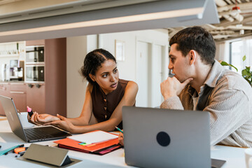 Female and male employees talking and sitting at a table in front of laptops while she holds a marker and he holds a pencil