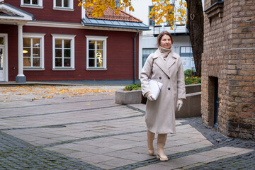 Elegant woman in a stylish beige coat walks confidently along a cobblestone path, surrounded by autumn leaves and charming architecture, embodying urban sophistication and seasonal beauty