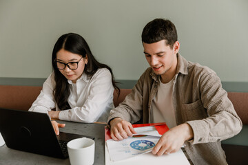 A female employee scrolls through a laptop and sits at a table next to a male employee who is smiling and putting a document in a folder