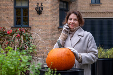 Woman in a cozy beige coat stands beside a large pumpkin in an outdoor garden, surrounded by vibrant plants, showcasing autumn's seasonal beauty and festive spirit