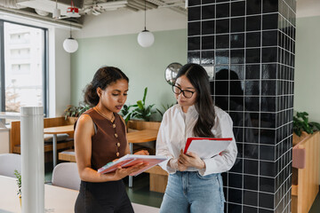 A female worker holds a folder and listens to a female worker who is talking and points to a notebook they are looking at while standing