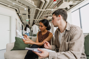 Fototapeta premium Female worker points with a marker at a document she is showing to a male worker holding a notebook while they sit on a couch