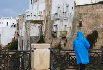 solitary figure in a bright blue raincoat stands on a railing overlooking the historic white and stone architecture of an italian town on a moody, overcast day.
