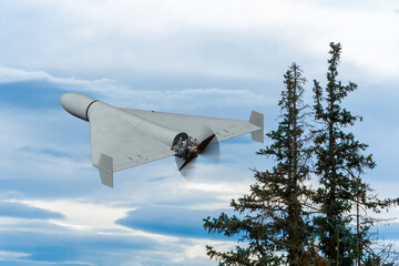 An unmanned aerial vehicle flies over the trees in a cloudy sky.
