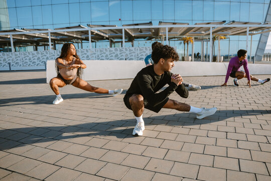 A group of four athletes squatting and looking to the side while stretching their legs and holding their hands together in front of them - Powered by Adobe