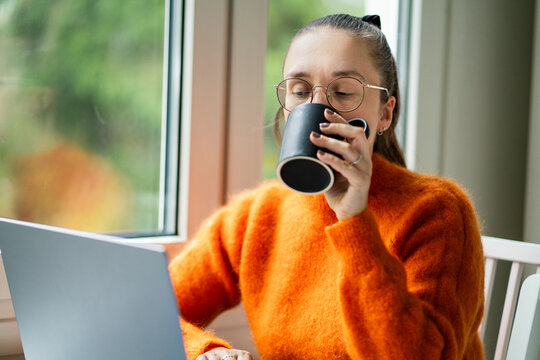 Woman drinking coffee while working from home. Cup of tea and laptop. Reading morning news and using computer. Busy person. - Powered by Adobe