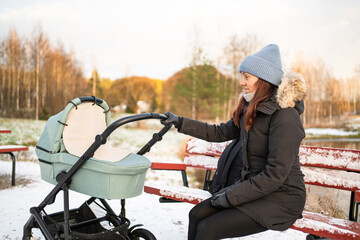 Stroller and mother in winter. Baby sleeping in carriage. Pram in snow. Tired woman sitting on park...