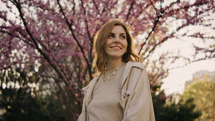 Woman stands under blooming pink trees in park during late afternoon while smiling and enjoying the scenery © tol_u4f