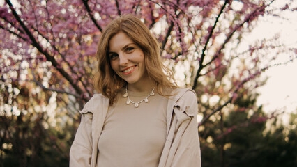 Woman stands in front of cherry blossom tree, smiling during sunny afternoon in spring, wearing...
