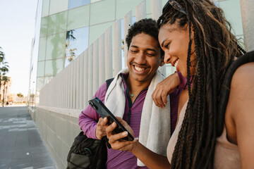 A male and female athlete stand and laugh while looking at a phone which she shows while he rests his hand on her shoulder