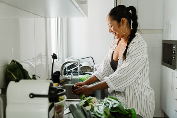 Woman laughing and washing cucumber in sink with tap water