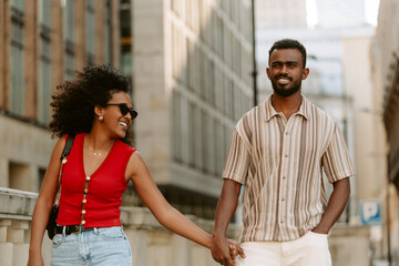 Woman laughing and listening to man talking and keeping hand in pocket as they hold hands and walk